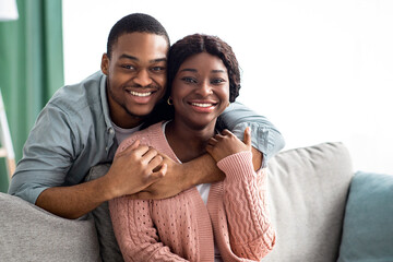 Portrait of happy black couple or husband and wife hugging at home, sitting on couch in living...