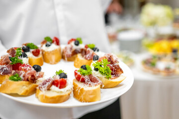Close-up of assorted appetizers on toasted bread with cherry tomatoes, olives, cheese, and cured meat, served on a white plate by a person in formal attire.