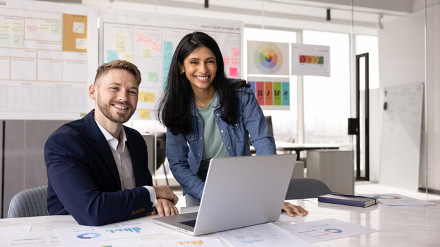 Diverse couple of happy millennial business colleagues meeting at office workplace with laptop, looking at camera, smiling, using computer for teamwork, standing and sitting for portrait together