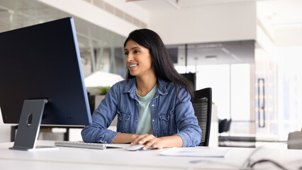 Positive young Indian professional woman in casual working on online project, Internet startup at computer monitor at workplace, sitting at office table in workspace alone, using mouse, smiling