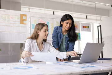 Two positive young multiethnic product managers working at laptop and paper marketing reports together, discussing online sales statistic benchmarks, watching content on computer