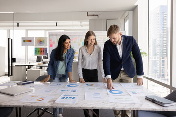 Diverse team of positive young coworkers in casual analyzing paper marketing reports on large office table, brainstorming on ideas for business plan, strategy, discussing sales analytics