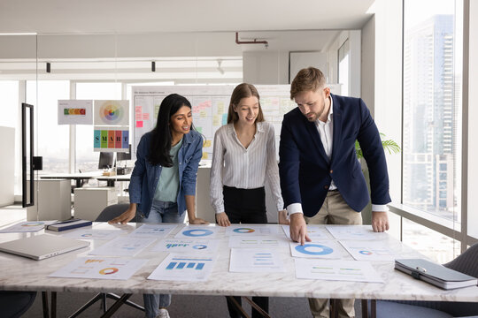 Diverse team of positive young coworkers in casual analyzing paper marketing reports on large office table, brainstorming on ideas for business plan, strategy, discussing sales analytics