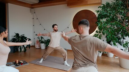 A male yoga mentor talks to a group of four students as they warm up with their arms out to the sides