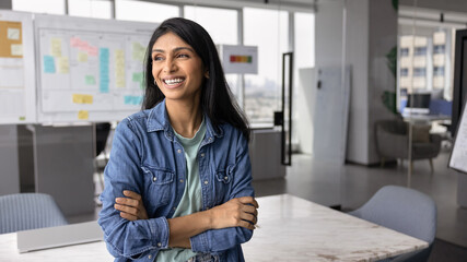 Happy successful young Indian startup owner woman in casual standing in office workspace with hands folded, looking away, smiling, laughing, thinking on successful project. Banner shot