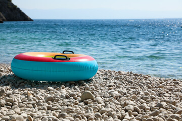 Fototapeta premium Bright children's inflatable swim ring on a pebble beach by clear turquoise water, with rocky coastline and sunny sky, evoking a fun and relaxing summer day at the seaside.