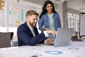 Multiethnic couple of cheerful business colleagues using laptop at workplace together, pointing at screen, smiling, laughing, meeting at large workplace table in collaboration room