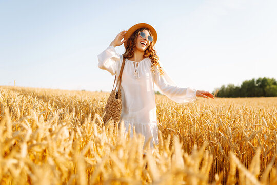 Beautiful woman in a hat walks in a golden wheat field at sunset. Happy woman in a white dress enjoys a rural landscape. Beauty, fashion, weekend concept. Lifestyle.
