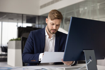 Fototapeta premium Serious young business company director man reviewing legal financial document at computer, checking invoice, bill. Professional analyst, expert working on paper report at office workplace