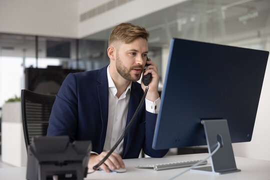 Positive busy young manager man in formal jacket talking on landline phone at office workplace, picking up call, using work application on desktop computer while giving telephone consultation