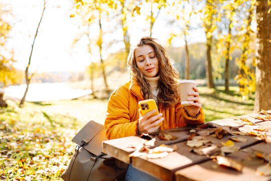 Сheerful woman in a bright jacket with phone and a glass of coffee sits at a wooden table in autumn park. A beautiful woman enjoys sunny weather and drinks hot drink. Weekend and drinks concept.