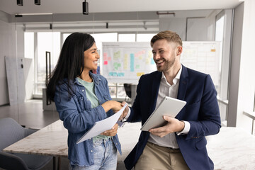 Positive multiethnic couple of coworkers shaking hands in office space, holding tablet, documents, smiling, laughing. Caucasian businessman thanking female Indian coworker for professional help