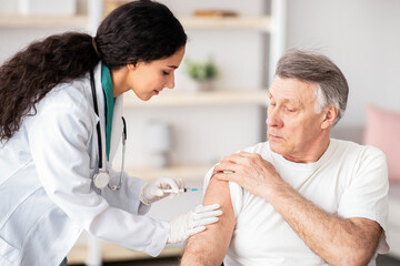 Obraz premium Young doctor making injection of medication or anti-covid vaccine to disabled elderly man in wheelchair at home. Medic immunizing senior handicapped patient, doing shot of antibiotic indoors