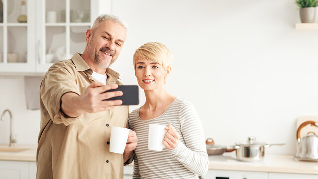 Self-isolation, video call, morning coffee. selfie at home and online meeting. Smiling middle aged caucasian man and woman with cups of hot drink makes selfie on smartphone in kitchen interior