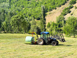 Obraz premium Tractor Baling Hay in a Mountain Field on a Summer Day