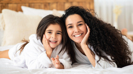 Adorable young mother and little daughter in bathrobes bonding on bed at home, happy mom and kid having beauty day, laying on bed in bedroom, hugging and cheerfully smiling at camera, copy space