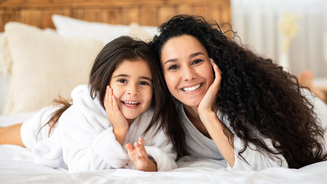 Adorable young mother and little daughter in bathrobes bonding on bed at home, happy mom and kid having beauty day, laying on bed in bedroom, hugging and cheerfully smiling at camera, copy space