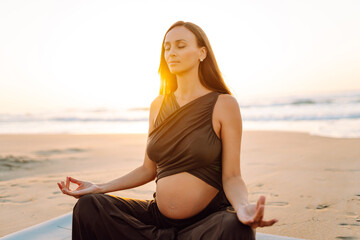 Pregnant woman with yoga mat practicing relaxation exercises on sandy beach. Beautiful expectant mother exercising and meditating at sunrise enjoying sea view. Health, relaxation concept.