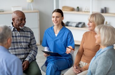 Friendly pretty young woman in blue workwear psychologist having conversation with multiracial group of senior people sitting in circle, psychological support for senior people at nursing home