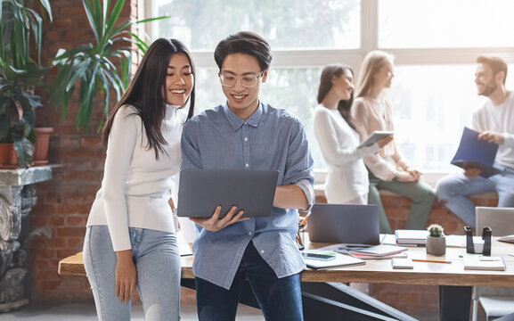 Excited asian guy sharing business ideas with young mixed race woman coworker, man and woman looking at laptop screen, office interior
