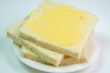 A close-up, angled shot of a stack of fresh white bread slices, with the top slice generously coated in a layer of yellow butter or margarine, on a simple white plate.