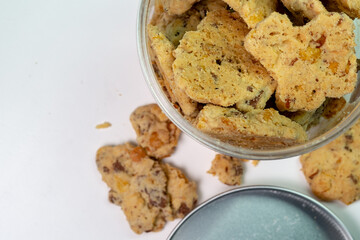 A top-down view of a clear plastic jar filled with delicious, crunchy almond cookies on a white background. Some cookies have spilled out, adding to the visual.
