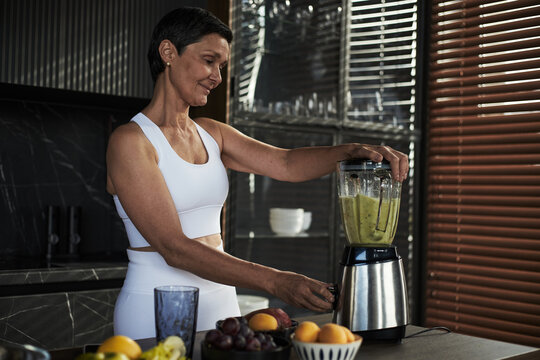 Middle aged Caucasian woman blending smoothie in kitchen, smiling while preparing healthy drink, standing near countertop with fresh fruits, wearing athletic clothing