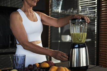Middle aged Caucasian woman blending green smoothie in kitchen, wearing sportswear, preparing healthy drink with fresh fruits visible on counter, focusing on healthy lifestyle