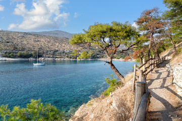 Scenic coastal pathway lined with trees, leading to a tranquil blue bay with a sailboat, surrounded by hills and clear skies, inviting outdoor exploration and relaxation. Greece, Thassos.