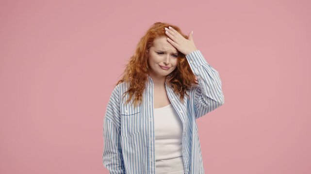 Frustrated red-haired woman sighs, puts hand on head, she forgot something. Facepalms after unexpected reaction on pink