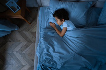 A young African American boy is laying comfortably in bed with a soft blue comforter covering him. He appears relaxed and at ease, possibly getting ready to sleep or just waking up, view above