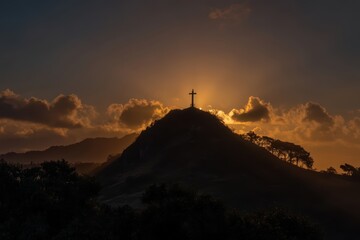 A cross stands atop a hill at sunset, surrounded by dramatic clouds and silhouetted trees against a glowing sky