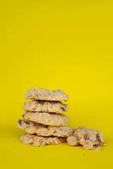A vertical close-up shot of a small stack of almond cookies, with some broken pieces to the side, all on a vibrant yellow background. The cookies show a crunchy texture with embedded nuts.