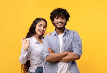 Obraz premium Portrait of happy indian couple embracing, woman showing okay gesture and smiling at camera over yellow studio background. Young hindu spouses approving or recommending something