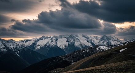 Dramatic mountain range under a stormy sky at sunset.  Snowy peaks pierce a dark, cloudy horizon.  Rolling hills slope downward