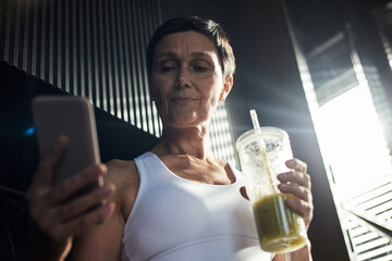 Middle aged Caucasian woman standing indoors holding smartphone in one hand and green smoothie in other hand, wearing athletic top, looking at phone screen