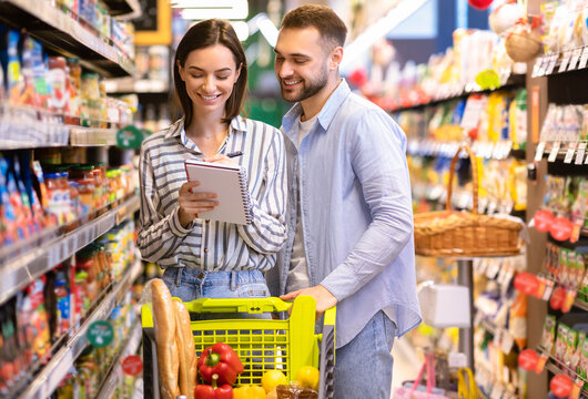 Portrait Of Two Happy Customers Holding Grocery Shopping Checklist, Writing In Notebook, Buying Products, Standing With Full Shop Cart In Supermarket. Young Millennial Family Choosing Healthy Food - Powered by Adobe