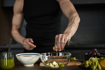 Caucasian man preparing healthy salad, squeezing lime over glass bowl filled with chopped fruit, muscular arms visible, kitchen counter with fresh ingredients