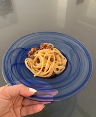 Woman holding a plate of square shaped spaghetti topped with a bolognese sauce in a glass blue bowl against a grey background 