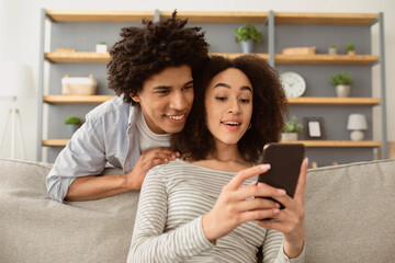 Interesting message, online call during COVID-19 pandemic. Cheerful excited millennial african american guy and lady look at smartphone and talk with family in cozy living room interior, copy space