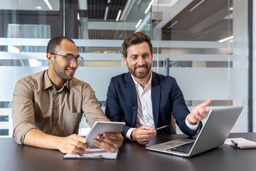Business colleagues working together on a laptop and tablet, collaborating on a project in a well-lit, modern office environment.