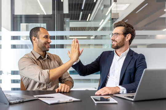Two coworkers share a high five while sitting at a table with laptops and paperwork in a bright office environment, symbolizing team collaboration, success, and workplace achievement.