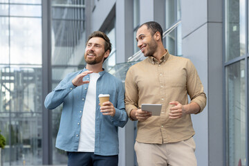Two friends engaging in a casual conversation outdoors, holding coffee and a tablet, within a contemporary cityscape environment.