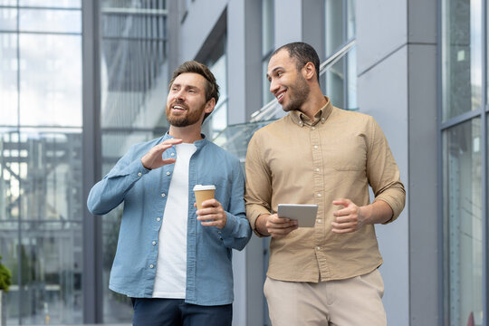 Two friends engaging in a casual conversation outdoors, holding coffee and a tablet, within a contemporary cityscape environment.