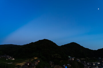 夕暮れ深く青の時間の山にかかる反薄明光線と月