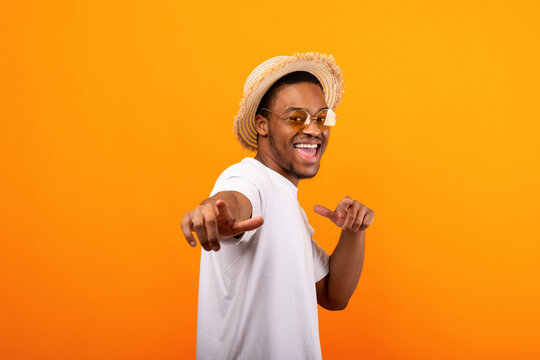 Happy black guy in casual summer outfit, straw hat and sunglasses pointing at you on yellow studio background. Positive African American man enjoying summertime, having fun