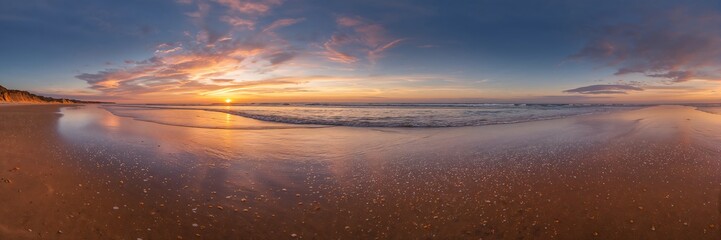 A panoramic view of a beach at sunset showing the reflection of the sky in the wet sand and ocean water