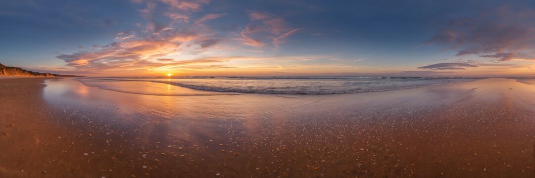 A panoramic view of a beach at sunset showing the reflection of the sky in the wet sand and ocean water - Powered by Adobe