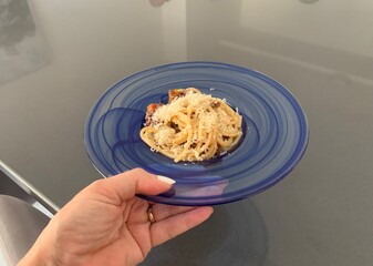 Woman holding a plate of square shaped spaghetti topped with a bolognese sauce and grated Parmesan cheese  in a glass blue bowl against a grey background 