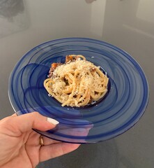 Woman holding a plate of square shaped spaghetti topped with a bolognese sauce and grated Parmesan cheese  in a glass blue bowl against a grey background